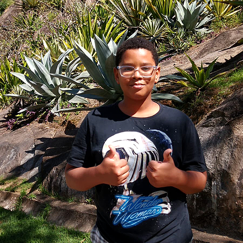 Menino de pele negra vestindo uma camiseta estampada e fazendo sinal de "joia" com as duas mãos. Ao fundo um parque arborizado.