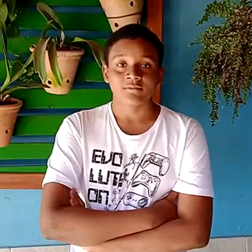 Adolescente de pele negra, cabelos curtos na cor preta, olha para a camera com os braços cruzados. Ao fundo diferentes plantas penduradas.