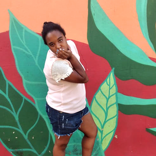 Flaviana, menina de pele negra e cabelos da cor preta. Ela veste uma blusa na cor branca e uma bermuda jeans.