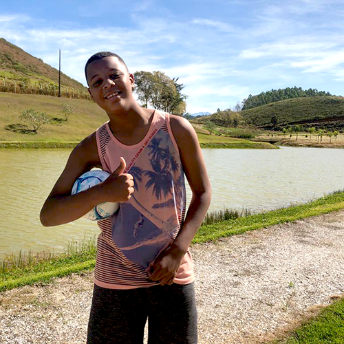 Salatiel, jovem de 13 anos de pele preta. Ele usa camiseta regata estampada e bermuda. Ele está em uma área rural e faz o sinal de positivo com a mão.