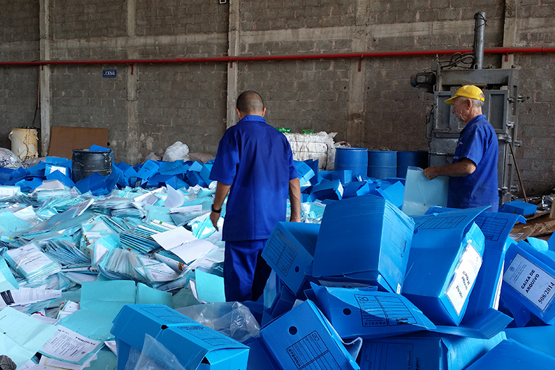 Funcionários de uma usina de reciclagem de papel caminham sobre pilha de caixas e processos que serão reciclados.