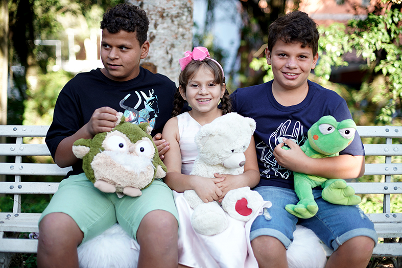 três crianças, uma menina e dois meninos, estão sentados em um banco de praça. Cada um está segurando um bicho de pelúcia.