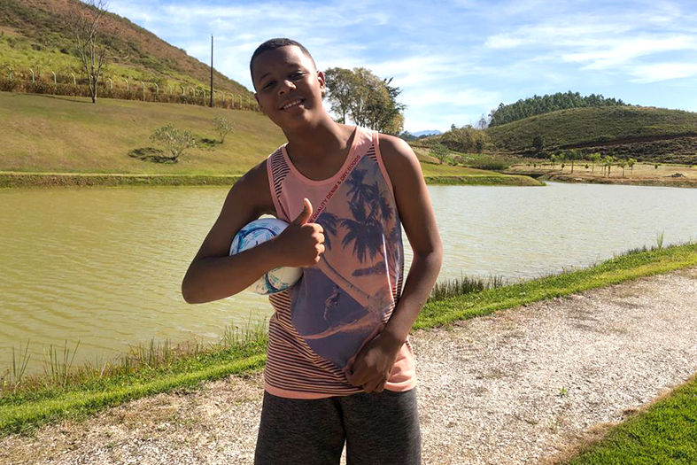 Salatiel, jovem de 13 anos de pele preta. Ele usa camiseta regata estampada e bermuda. Ele está em uma área rural e faz o sinal de positivo com a mão.