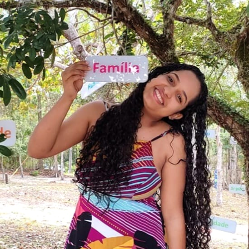 Fernanda, adolescente de pele parda e cabelos compridos e anelados na cor castanho escuro. Ela usa um vestido colorido e segura uma placa com a palavra "família" gravada.