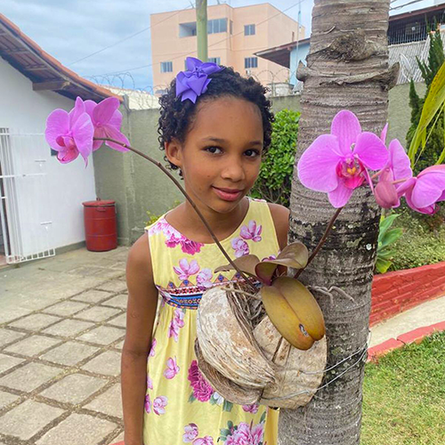 A menina Kezia Adriani de 9 anos tem pele parda, os cabelos castanhos e encaracolados. Ela veste um vestido amarelo com detalhes em outras cores.