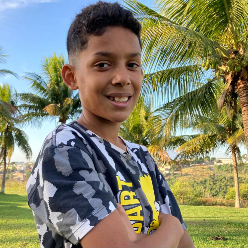 menino de pele negra e cabelos pretos. ele está usando uma camisa estampada. ao fundo vemos um parque com coqueiros.