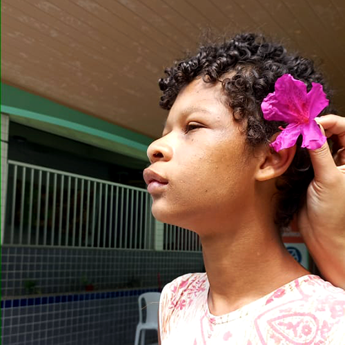 Menina de pele na cor preta, cabelos na cor preta, posa de perfil para a câmera. Em seu cabelo uma outra pessoa coloca uma flor de cor violeta.