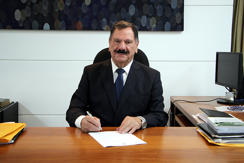 presidente do tjes, desembargador ronaldo gonçalves de souza sentado em frente a uma mesa com tampo em madeira. com a mão esquerda ele segura uma folha de papel, com a esquerda ele toca o papel com uma caneta. ele olha para a câmera.