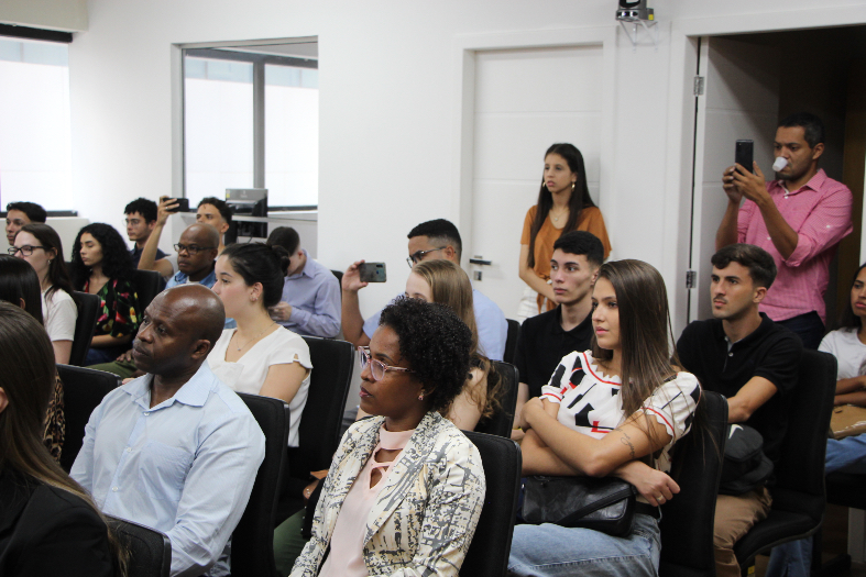 Pessoas sentadas nas cadeiras da sala de sessões assistem uma palestra.
