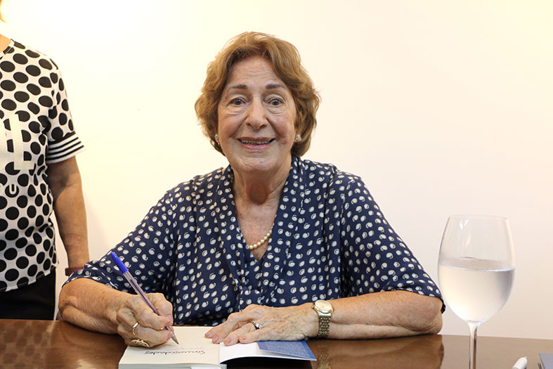 A escritora Terezinha Tristão Bichara, mulher de pele branca vestindo um vestido azul decorado com bolinhas brancas, posa para a foto olhando para a câmera e sorrindo.