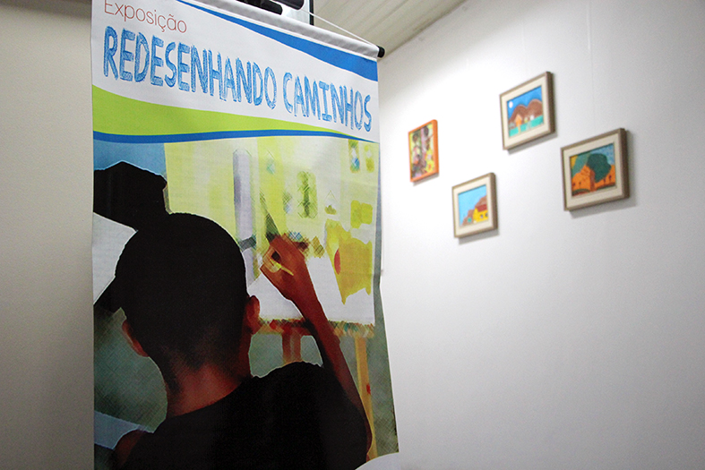 Em primeiro plano banner com a arte da exposição dos adolescente do IASES em segundo plano e desfocado 4 quadros pequenos pintados à mão