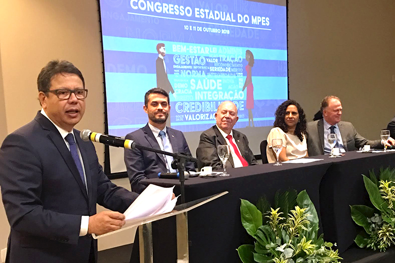 Um homem discursa em um púlpito enquanto outras 4 pessoas observam sentados próximos a ele em uma mesa de honra.