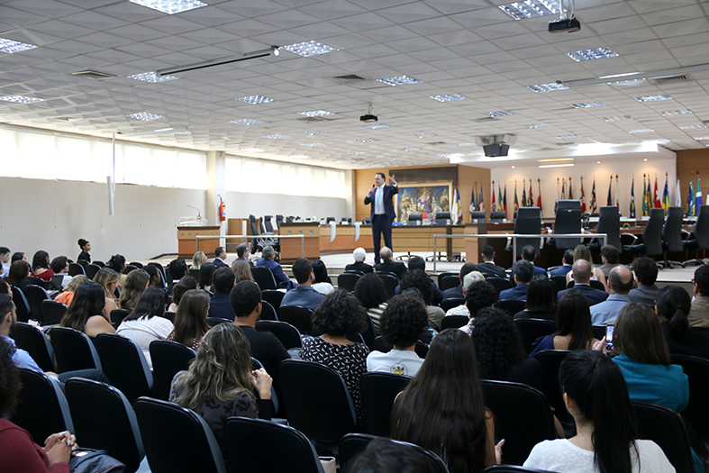 Auditório (Salão do Pleno) cheio de pessoas assistindo a um homem de terno falar de pé ao microfone.