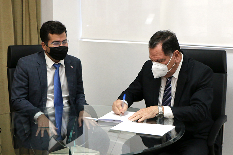 Dois homens sentados em frente a uma mesa circular com tampo de vidro. Um deles assina um documento enquanto o outro o observa.