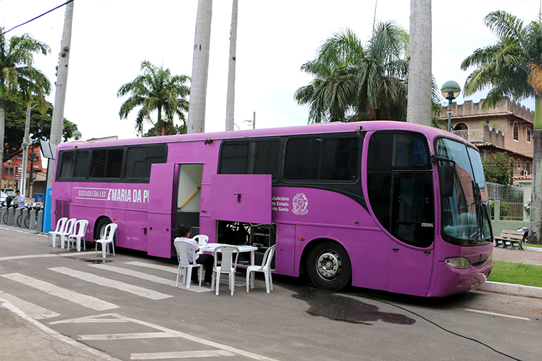Ônibus do Juizado Itinerante da Lei Maria da Penha estacionado, com as portas abertas e uma mesa com quatro cadeiras postas do lado de fora.