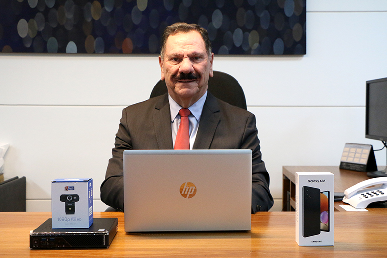 O presidente do TJES, Desembargador Ronaldo Gonçalves de Sousa, homem de pele branca e cabelos e bigode na cor preta olha para a câmera. Em cima da mesa estão diversos equipamentos eletrônicos como smartphone, webcam e notebook.