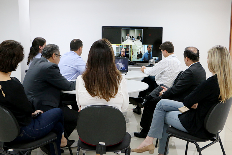 Pessoas sentadas em caderias em torno de uma mesa redonda, de costas para a câmera, assistem a uma videoconferência