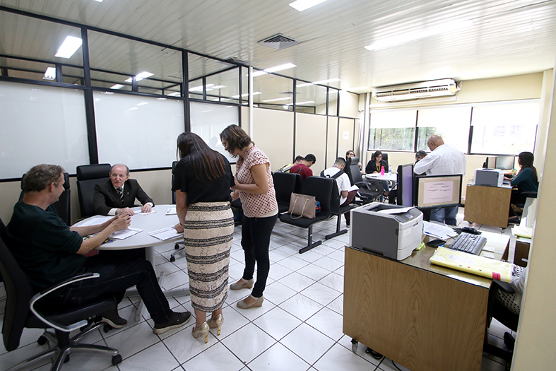 Pessoas trabalhando entre em pé e sentadas em uma sala com divisórias de vidro e madeira compensada