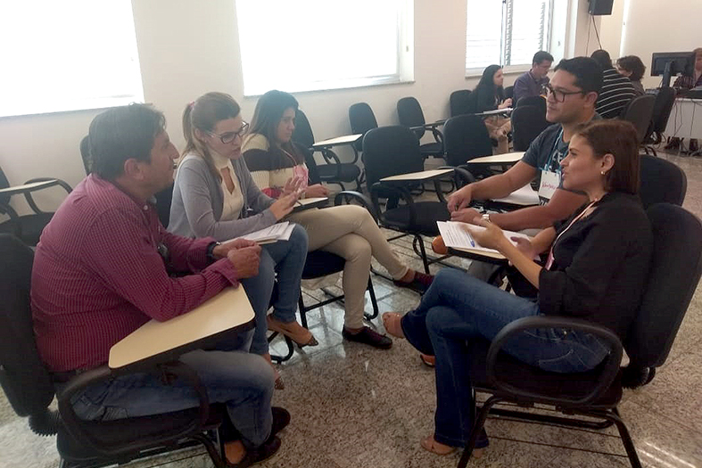 Grupo de pessoas sentadas em círculos de cadeiras do tipo carteira escolar.