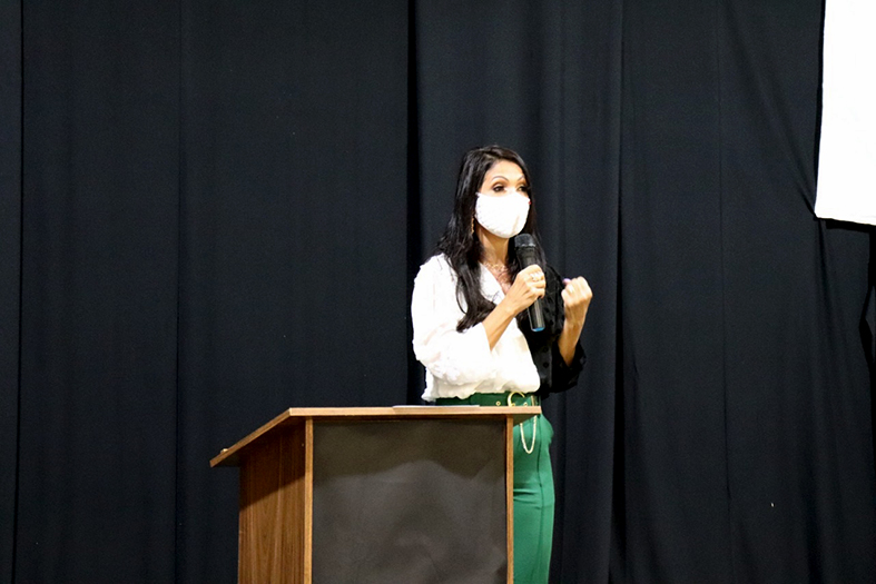 uma mulher de pele branca, de estatura mediana, com cabelos pretos e vestindo saia verde e camisa branca fala em um microfone. Frente à ela está um púlpito de madeira e, ao fundo, uma cortina na cor preta.