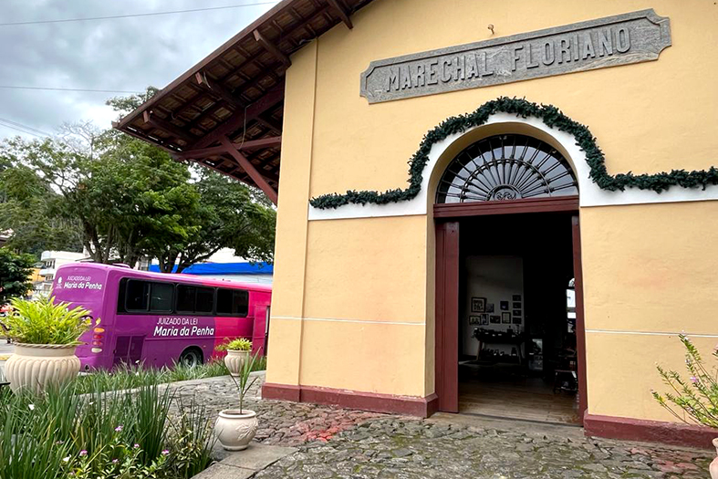 Ônibus do Juizado Itinerante da Lei Maria da Penha estacionado próximo a uma antiga estação de trem onde lê-se na fachada do prédio os dizeres "Marechal Floriano".