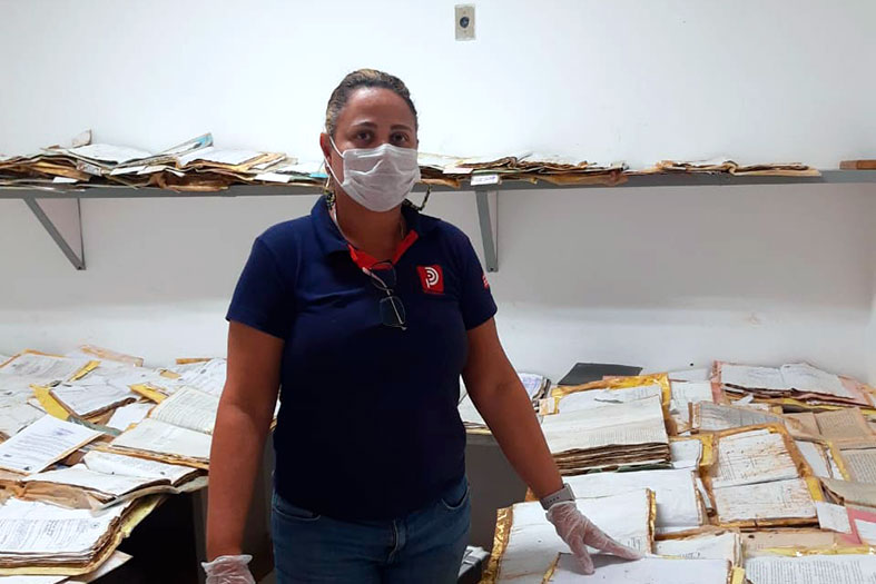 Colaboradora da empresa de restauração de processos posa para foto em uma sala cheia de processos. Ela usa camisa azul marinho, calça jeans, máscara de proteção no rosto e touca higiênica na cabeça.