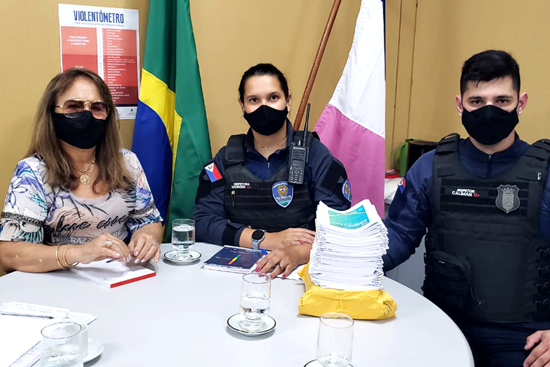 juíza coordenadora da coordenadoria de combate à violência doméstica e familiar drª hermínia azoury reunida em uma mesa redonda com um guarda e uma guarda municipal da cidade de vila velha. em cima da mesa estão dispostos materiais de campanhas contra à violência doméstica.