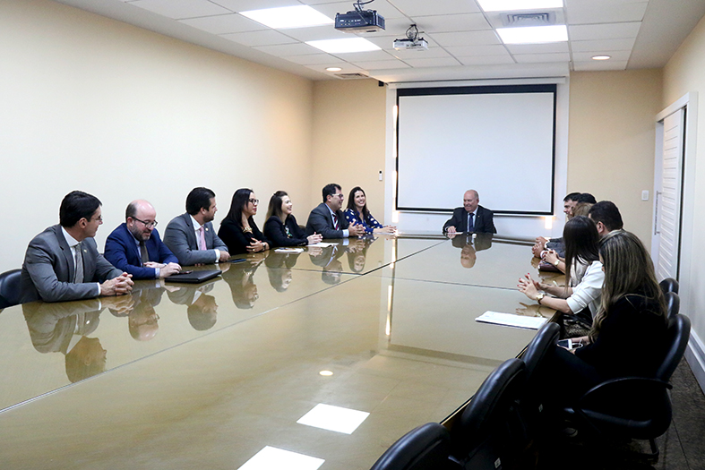 Grupo de pessoas reunidas em torno de uma grande mesa ovalada e de tampo de vidro.