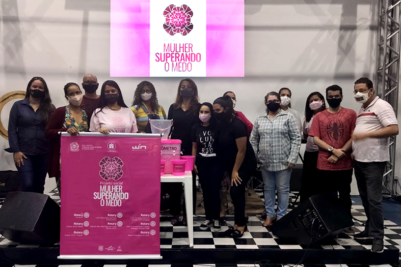 grupo de pessoas posa para foto num palco. A frente do grupo um banner de cor rosa com os dizeres "Projeto Social - Mulher Superando o medo". Ao fundo do palco a mesma imagem é projetada em um telão.