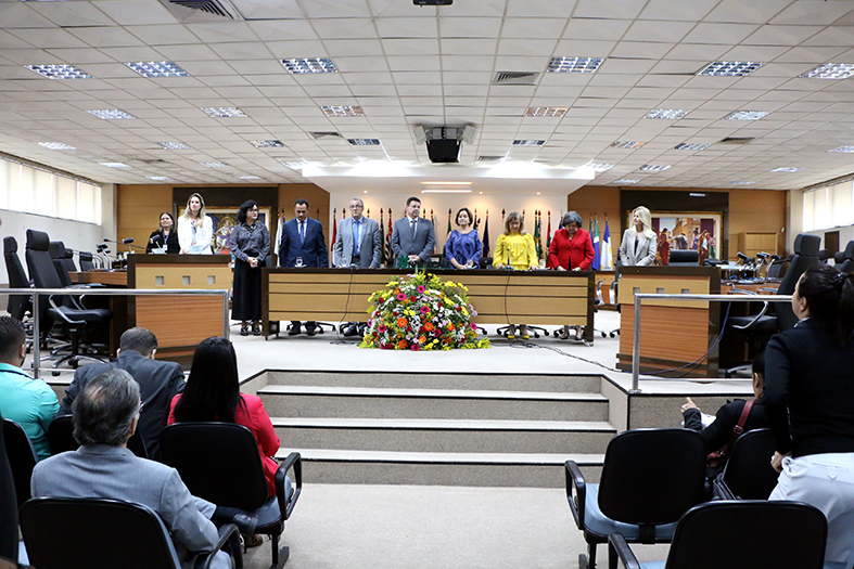 Grupo de pessoas com trajes formais se encontram de pé e perfiladas em respeito ao hino nacional. todas estão atrás de uma mesa decorada com flores dentro de um grande auditório.