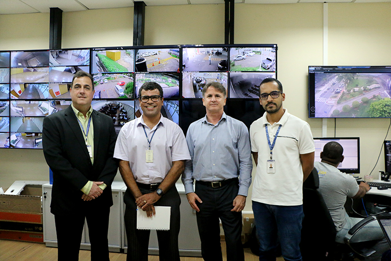 Quatro homens, 1 de terno e outros três de camisa social posam para foto. Ao fundo um painel de telões.