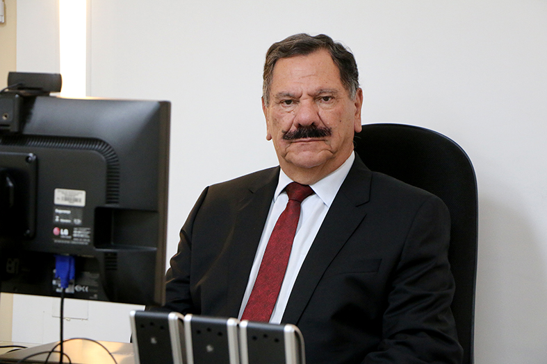 Homem branco com cabelo e bigode pretos, de terno e gravata, sentado e com a parte traseira de um monitor em primeiro plano olha para a câmera.