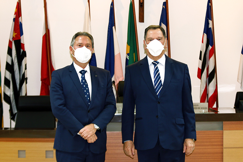dois homens brancos posam lado a lado para foto. eles estão suando máscara na cor branca e ternos na cor azul marinho.