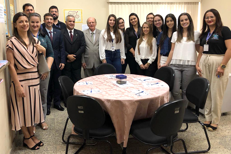 Grupo de pessoas em pé posando para a foto em trono de uma messa redonda com papéis e canetas em cima.