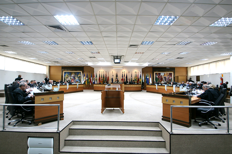 Foto panorâmica do salão pleno do tribunal de justiça onde se vê 30 assentos e ao fundo baneidras dos estados e dois quadros bem coloridos.