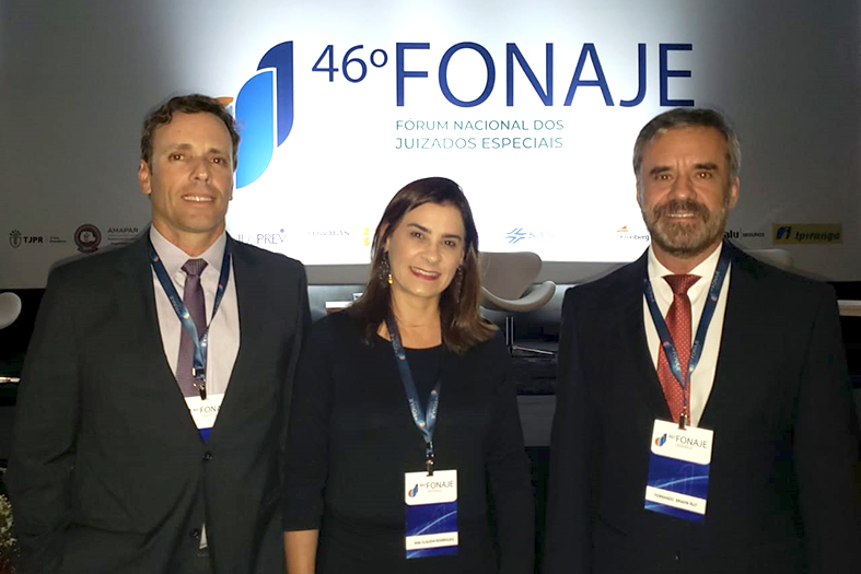 Dois homens de terno e uma mulher de vestido em meio à eles possam para foto em frente a uma projeção onde lê-se 46º Fonaje