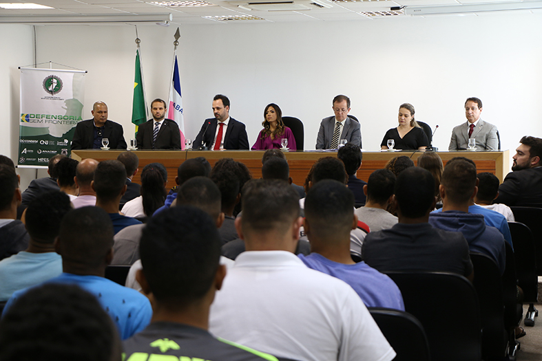 Várias pessoas dentro de um auditório assistindo a uma audiência onde 6 pessoas estão no palco.