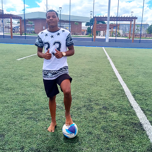 Menino de pele negra e cabelo preto e curto. Ele veste camiseta e bermuda e está com um dos pés encima de uma bola de futebol.