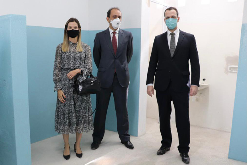 três pessoas, dois homens brancos e uma mulher branca, posam para fotos. eles estão usando terno na cor preta e ela está usando vestido longo e estampado.