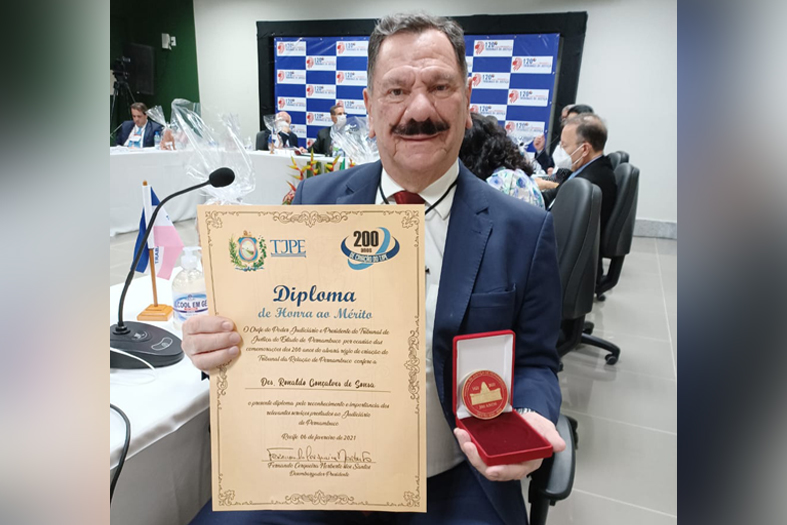 O presidente do TJES, Desembargador Ronaldo Gonçalves, home de pele branca e de estatura alta, com cabelos e bigode preto, posa para foto segurando uma medalha e um diploma.