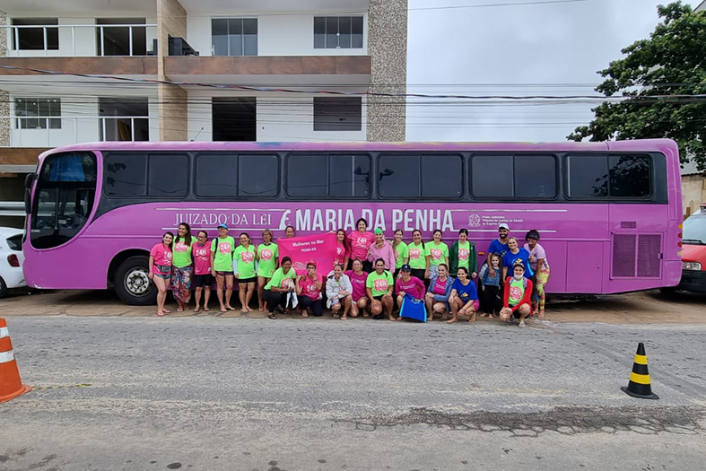 Grupo grande de pessoas posa para fotografia em frente ao um ônibus tipo leito convertido em escritório itinerante, O ônibus está pintado da cor rosa.