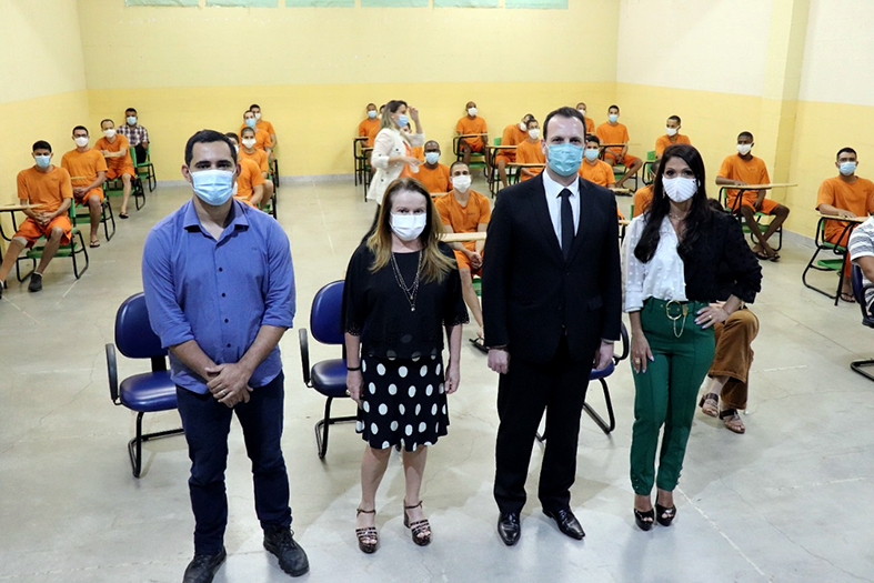 quatro pessoas de pele branca posam para a foto, sendo duas mulheres e dois homens. Eles estão de pé e, ao fundo, sentados em carteiras afastadas estão vários internos do sistema prisional. Todos na foto estão de máscara.