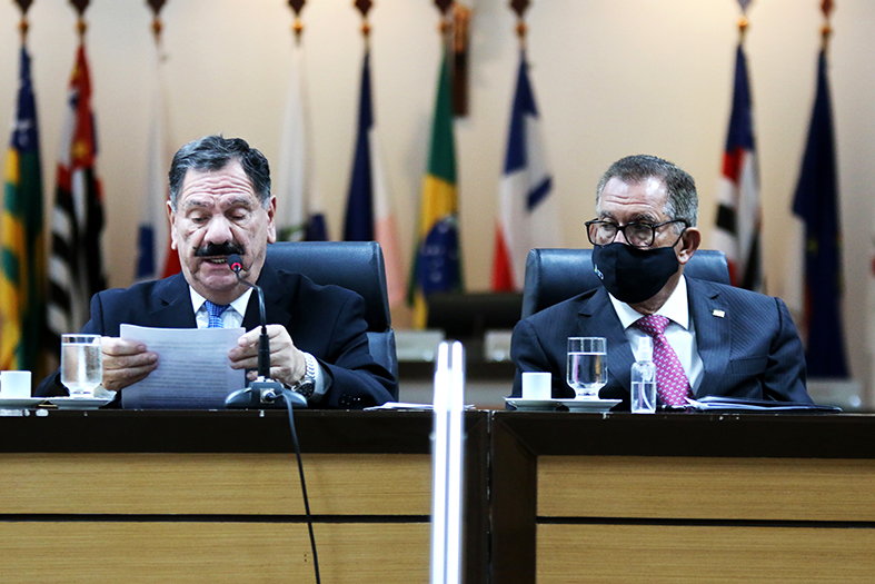 O presidente do TJES, Desembargador Ronaldo Gonçalves, um homem de pele branca e de cabelos e barba da cor preta está falando ao microfone. Ao seu lado, usando máscara, o ministro do STJ, Humberto Martins, um homem de pele morena e de cabelos grisalhos o observa.