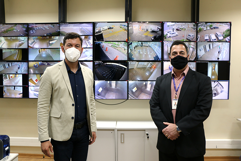 Dois homens posam para foto. Ao fundo uma parede de televisores com imagens de câmeras de segurança.