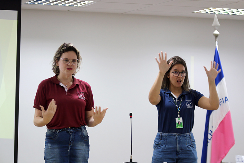 Duas mulheres falam ao público durante homenagem, uma delas é intérprete de libras.