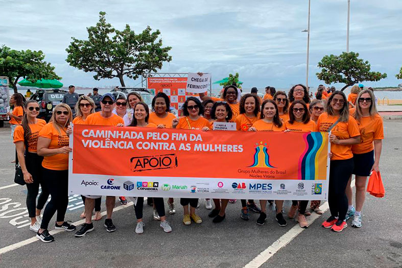 Um grupo de mulheres vestindo camisas de cor laranja posa para foto com uma faixa da mesma cor com os dizeres "caminhada pelo fim da violência contra as mulheres"