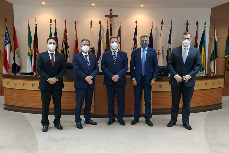 grupo de 4 homens brancos posam para foto lado a lado usando máscaras. eles estão todos trajando ternos. ao fundo um amplo salão decorado com bandeiras de estados do brasil.