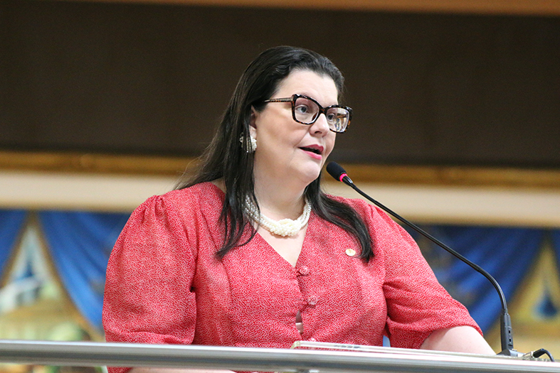 A procuradora Luciana Andrade, mulher de pele branca, cabelos na cor preta e óculos com armação preta, fala ao microfone.