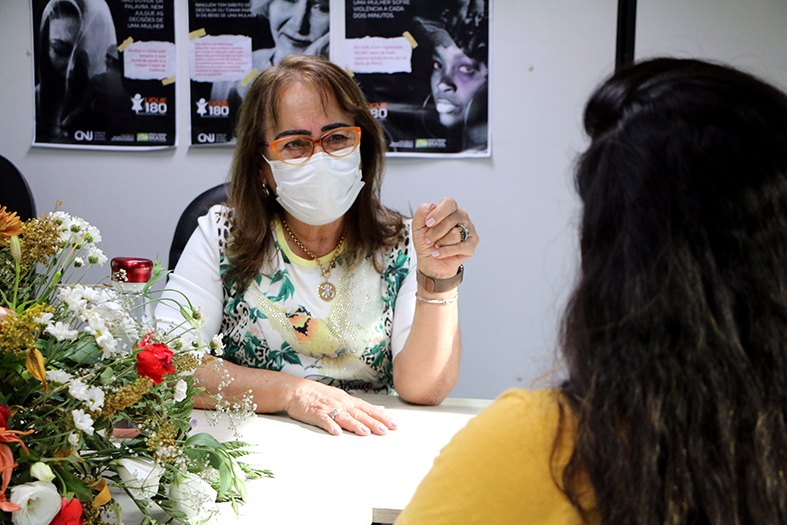 Drª Hermínia Azoury, mulher de pele branca, cabelos tingidos de loiro, usando vestido branco conversa com uma mulher de cabelos castanhos de costas para a foto.