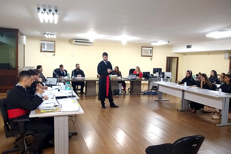 EM um salão amplo um jovem vestindo toga de magistrado se apresenta para um grupo de pessoas sentadas em cadeiras em torno dele.